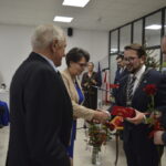 Na zdjęciu kilku elegancko ubranych ludzi, trzymają kwiaty, wręczając nagrody w jasno oświetlonym pomieszczeniu. Obok choinka. Na ścianie flagi.