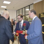 Na zdjęciu widnieje grupa osób w eleganckich strojach podczas uroczystości. Tło jest jasne z dekoracją ZŁOTE GODY i kwiatami. Osoby wymieniają uściski dłoni i wręczają róże.