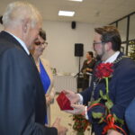 Na zdjęciu widać grupę ludzi podczas uroczystości. Mężczyzna w garniturze wręcza czerwoną różę i czerwone pudełko. W tle choinka i stoły z jedzeniem.