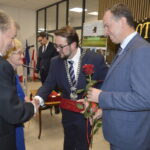 Grupa osób w garniturach, mężczyzna z łańcuchem trzyma różę. W tle flagi i dekoracyjna tablica. Formalna atmosfera.