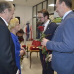 Na zdjęciu cztery osoby w formalnych strojach wymieniają się różami w jasnym pomieszczeniu. Tło zdobią flagi i czarny plakat z napisem. Atmosfera jest uroczysta.