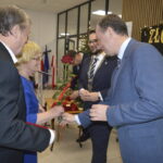 Grupa osób ubranych elegancko, mężczyzna wręcza kobiecie różę. Tło jasne, napisy „Złote” w tle. Widać flagi i mikrofony.