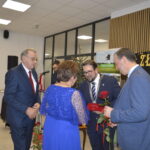 Grupa ludzi w eleganckich strojach stoi w sali. Mężczyzna wręcza kobiecie różę i upominek. Tło z napisem Złote.