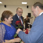 Starsza kobieta w niebieskiej sukience przyjmuje czerwoną różę od mężczyzny w szarym garniturze, obok stoi starszy mężczyzna.