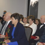 Grupa elegancko ubranych osób siedzi w sali, trzymając czerwone róże i kubki z napisami. Tło jest jasne i neutralne. Event w formalnym otoczeniu.