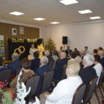 Sala z ludźmi na uroczystości Złote Gody. Występująca osoba na tle dekoracji z złotymi obrączkami. Świerki z lampkami.