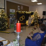 To zdjęcie z przyjęcia „Złote Gody”. Na pierwszym planie czerwono ubrana świeca, w tle choinki z lampkami. Goście siedzą przy stole, para obrączek na czarnym tle.