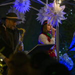 Muzycy w strojach góralskich grają na scenie oświetlonej lampkami. W tle białe dekoracje przypominające gwiazdy. Dominuje ciemne tło z kolorowymi światłami.