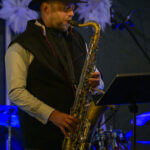 Muzyk w czarnym kapeluszu gra na saksofonie. Tło ciemne z białymi ozdobami. Scena oświetlona na niebiesko.