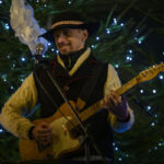 Muzyk w kapeluszu gra na gitarze elektrycznej. W tle udekorowana choinka z niebieskimi światełkami. Sceneria nocna, świąteczna atmosfera.