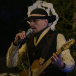 Mężczyzna w kapeluszu gra na gitarze i śpiewa do mikrofonu. Ma na sobie jasny sweter i ciemną kamizelkę. W tle ciemne drzewo.