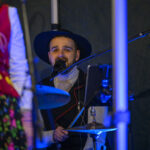 Muzyk gra na perkusji i śpiewa do mikrofonu. Nosi kapelusz i jasny sweter na ciemnym tle. Obok widoczne są dekoracje i mikrofon.