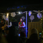 Na scenie pod namiotem występuje zespół w tradycyjnych strojach. W tle zielone choinki i białe dekoracje. Wokół delikatne światełka.