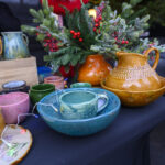 Na czarnym stole stoją kolorowe, ceramiczne naczynia i zielona kompozycja z czerwonymi jagodami. Obok znajduje się drewniana skrzynka. W tle widać osobę w ciemnym ubraniu.