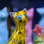 Kolorowe ceramiczne figurki kotów na kolorowym, rozmytym tle. Żółty kot z niebieskimi oczami na pierwszym planie. Barwy tła: niebieskie i różowe.