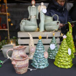 Kolorowe ceramiczne ozdoby i naczynia na czarnym stole. W tle widać osoby oraz półki z lampkami. Dominują zielenie i pomarańcze.