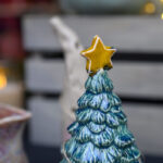 Ceramiczna choinka z zielonymi gałązkami i żółtą gwiazdą na szczycie. W tle rozmyte światła i neutralne tło. Dekoracja nawiązuje do świąt.