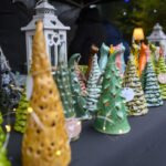 Na stole stoją kolorowe, ceramiczne choinki oraz latarnia. Tło jest ciemne, co podkreśla detale ozdób. Widać także lampki dekoracyjne.