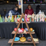 Stragan z ceramiką na tle czarnej tkaniny. Kolorowe figurki, misy i ozdoby choinkowe. Osoby ubrane w ciepłe kurtki.