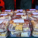 Plastikowe opakowania z różnorodnymi ciastkami na stole. W tle widoczne osoby w zielonych fartuchach. Ciepłe oświetlenie wnętrza.