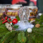 Na obrazie widoczna jest szklana figurka anioła ustawiona na zielonej choince. Ozdoby w kolorach czerwonym, białym i naturalnym dodają uroku. W tle pudełka z ciastkami tworzą przytulny klimat.