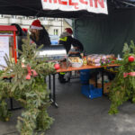 Stragan na jarmarku świątecznym z dekoracjami z gałązek iglastych. Czerwone i zielone akcenty, ludzie w czapkach Mikołaja. W tle ciemny namiot.