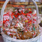 Koszyk wypełniony świątecznymi ciastkami w przezroczystych opakowaniach z czerwonymi wstążkami. Dominują kolory białe i czerwone. W tle rustykalny motyw dekoracyjny.