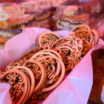 Drewniane ozdoby w koszu, na różowym tle. Obok słoiki i ciasta w pudełkach. Jasne kolory, świąteczny klimat.