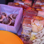 Na zdjęciu widać ciastka w kolorowych opakowaniach. Dominują barwy różowe i pomarańczowe. W tle znajduje się dekoracja choinkowa.