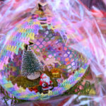 Kolorowy koszyk z ozdobą choinkową, Mikołajem i prezentami. Dominują barwy różowe, żółte i zielone. Tło rozmyte, przeźroczysta folia.