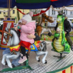 Karuzela z dziećmi siedzącymi na plastikowych zwierzętach. Dziewczynka w różowym stroju na koniu, chłopiec na zielonym smoku. W tle widać kolorowe budynki i drzewka.