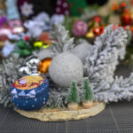 Dekoracja świąteczna z gałązkami, bombkami i miniaturowymi drzewkami. Dominują szarości i zielenie. Tło jest rozmyte z kolorowymi ozdobami.