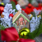 Domek z piernika z czerwonymi i zielonymi detalami wśród śnieżnych dekoracji. Tło to żywe kolory, głównie czerwienie. Obok bombki, wstążki i gałązki.