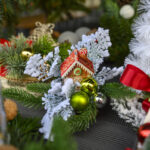 Świąteczna kompozycja z zielonymi gałązkami, białym śniegiem i kolorowymi bombkami. W centrum jest domek z piernika. Tło zdobią czerwone wstążki i dekoracje.