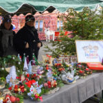 Stragan bożonarodzeniowy z wieńcami i ozdobami, tło z ceglaną ścianą i choinką. Dominują zieleń, czerwień i światełka. Dwie osoby w czapkach zimowych.