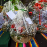 Kosze świąteczne zapakowane w folię, ozdobione szyszkami i czerwonymi kokardkami na kolorowym, pasiastym materiale. Tło stanowią słoiki i białe pudełka z napisami. Dominuje kolor zielony i czerwony.