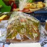 Ciasteczko piernikowe z kolorowymi dekoracjami, zapakowane w przezroczystą folię na świątecznym stole. W tle widać więcej wypieków i zielone ozdoby. Scena pełna kolorów i świątecznego klimatu.