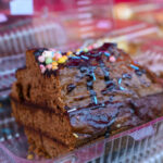 Kawałek ciasta czekoladowego z polewą i kolorową posypką w plastikowym pojemniku. Tło rozmyte, ciepłe odcienie.