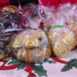 Doceniam Twoje pytanie! Zdjęcie przedstawia zapakowane w przezroczyste folie ciastka i przekąski, związane kolorowymi wstążkami. Produkty leżą na obrusie z motywem świątecznym w zielone liście i czerwone wstążki.