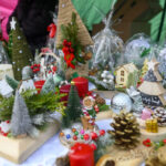 Świąteczny stół z ozdobami: czerwone świece, zielone choinki, złote szyszki, mini domki i bombki ośnieżone na białym tle.