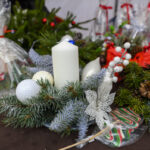 Wianek świąteczny z białą świecą, bombkami i gałązkami iglastymi na brązowym tle. Obok kolorowe ozdoby, w tym laski cukrowe. Tło zawiera zapakowane prezenty.