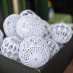 Białe, ażurowe kulki leżą w czarnym pudełku. W tle niewyraźne, zielono-brązowe tło. Kulki mają wzory przypominające koronkę.