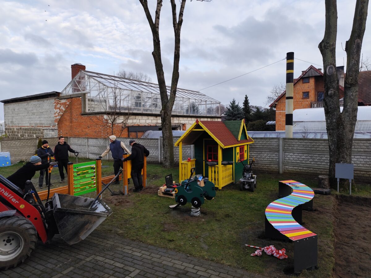 Plac zabaw z kolorową altanką, tęczową ławką i sprzętem do ćwiczeń. W tle osoby pracują i budynek z cegieł. Niebo pochmurne.