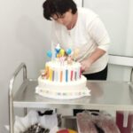 Kobieta w białej bluzce ustawiająca kolorowy tort na wózku. Tło jest jasne z białymi drzwiami. Tort ma dekoracje z balonami i liczbą 7.