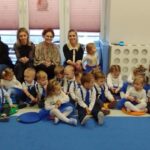 Grupa małych dzieci i cztery kobiety siedzą na dywanie. Dzieci mają eleganckie stroje, dziewczynki w niebieskich spódniczkach. Tło jest jasne, z zabawkami po lewej stronie.