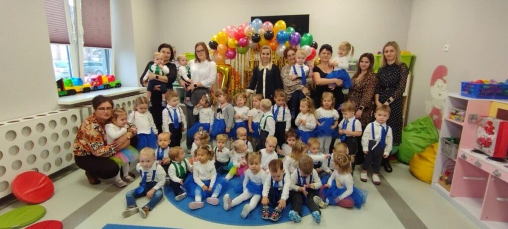 Na zdjęciu widoczna jest grupa dzieci i dorosłych w sali, ubrani w kolory niebieski i biały. Tło zdobi kolorowa girlanda z balonami. Widać również zabawki i dekoracje na ścianach.