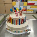 Tort z figurkami niebieskiego i beżowego stworka, kolorowe świeczki, cyfra 7 na świeczce. Tło: kolorowe szafki.