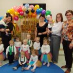 Grupa dzieci w kolorowych strojach z opiekunami stoi na niebieskiej macie. W tle balony i szare ściany. Obok zabawki i pluszowy Gargamel.