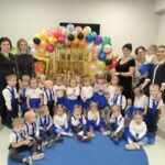 Grupa dzieci i nauczycielki w kolorowych strojach na tle balonów w sali. Dzieci mają niebieskie elementy ubrań. Pomieszczenie zdobi postać smerfa.