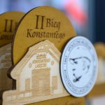 DSC_5348 Trofeum z drewna z napisem II Bieg Konstantego i medalem z twarzą na tle. Kolory beżowy i biały, rozmyte tło.